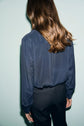 A person with long brown hair, wearing a bleu foncé blouse en soie with a delicate col volanté and black pants, stands facing away from the camera against a light-colored background.