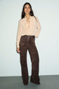 A woman stands against a light background, wearing a blouse femme with col volanté in beige, tucked into high-waisted, wide-leg brown suede pants with a belt. She has long dark hair and a relaxed pose.