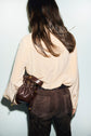 A person with long brown hair, wearing a beige blouse en soie and dark brown pants, stands with their back to the camera, holding a small brown leather drawstring bag.