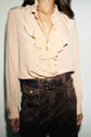 A person wears a pale col volanté blouse en soie with a gold necklace, paired with high-waisted brown pants and a woven belt with a gold buckle. The image is cropped from the chin to the upper thighs.
