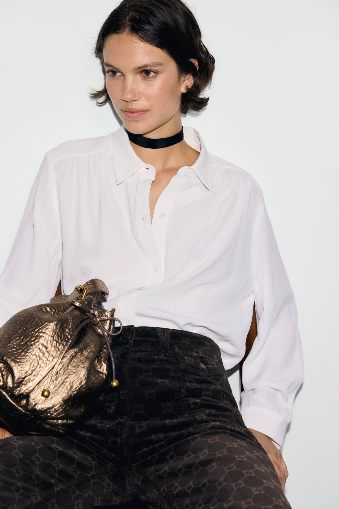 A person wearing a chemise écru button-up shirt, dark patterned pants, and a black choker sits holding a metallic gold textured bag. The background is plain white.