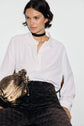A person wearing a chemise écru button-up shirt, dark patterned pants, and a black choker sits holding a metallic gold textured bag. The background is plain white.
