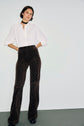 A woman stands against a plain wall, wearing an elegant chemise femme—a crisp white button-up shirt—paired with high-waisted dark brown patterned pants, black platform shoes, and a sleek black choker. She has short brown hair and assumes a relaxed posture.