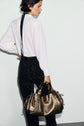 A woman in a chemise soie and dark pants stands sideways, holding a metallic gold handbag by her side against a plain light background.