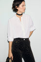 Woman wearing an elegant chemise écru, paired with black patterned high-waisted pants and a black choker. She has short dark hair and is holding a small bag, standing against a plain light background.