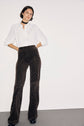 A woman stands against a white wall wearing a chic chemise femme—an elegant white button-up shirt—paired with dark high-waisted patterned pants, black shoes, and a black choker. Her hands are in her pockets and she looks confidently at the camera.