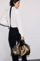 A person wearing a chemise écru and patterned black pants stands sideways, holding a metallic gold handbag, with a black ribbon tied at the back of their neck.