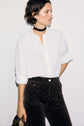 A woman wearing a chemise femme, specifically an écru white button-up shirt, dark patterned pants, and a black choker stands against a plain white background, holding a purse in her left hand.