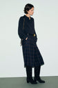 A woman wearing a black chemise en soie, plaid midi skirt, black knee-high boots, and a black belt with a gold buckle stands against a plain light background, hands in her pockets, with a shoulder bag—a perfect look classique chic.