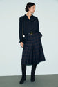Person wearing a black blouse reminiscent of a chemise en soie, dark plaid culottes, a black belt, black knee-high boots, and a black shoulder bag stands against a plain light background, perfectly embodying a look classique chic.