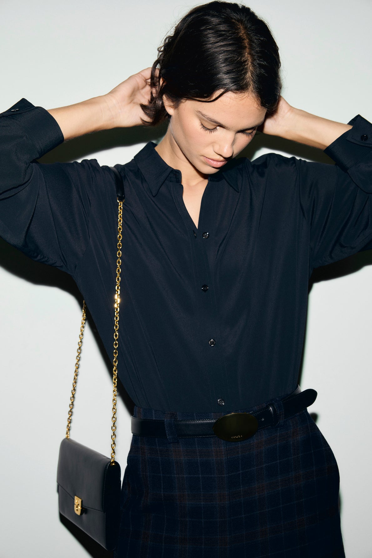 A person wearing a dark button-up shirt reminiscent of a chemise Lilie, paired with a plaid skirt and black belt, looks down, adjusting their hair. Their look classique chic is completed by a black purse with a gold chain strap over their shoulder.