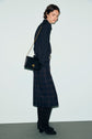 Woman wearing a chemise Lilie, plaid midi skirt, black knee-high boots, and holding a black shoulder bag with a gold chain, standing against a plain light background for a look classique chic.