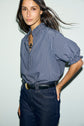 A woman with long brown hair wears a blouse rayée bleu et blanc à col montant à volants with dark high-waisted jeans and a black belt, standing against a plain light background.