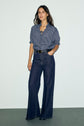 Woman standing against a plain wall, wearing a blouse rayée with manches courtes and a col montant à volants, tucked into high-waisted wide-leg dark blue jeans, accessorized with a black belt and necklace.