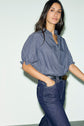 A woman with long dark hair wears a blue and white blouse rayée à col montant à volants, paired with dark blue jeans and a black belt, standing against a plain light-colored background.
