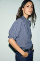 A woman with long brown hair wears a blue and white blouse rayée à col montant à volants with manches courtes and dark jeans, standing against a light background and looking at the camera.
