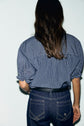 A person with long brown hair, seen from behind, wears a blouse rayée in blue and white with rolled sleeves and dark blue jeans featuring white stitching.