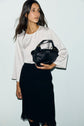A woman in a coupe trapèze black skirt and a top satiné écru with a délicat col perlé doré holds a black handbag against a plain light background.