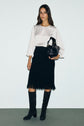 Woman standing against a plain background wearing a top satiné écru with a col perlé doré, paired with a black textured coupe trapèze knee-length skirt, black knee-high boots, and holding a black handbag.