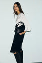 A woman stands against a light background, wearing a top satiné écru and a black skirt coupe trapèze with a fringed hem, paired with black knee-high boots. She holds a black handbag under her arm and looks toward the camera with a neutral expression.