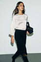 A woman with long brown hair wears a top satiné écru with a col perlé doré, styled elegantly with a black skirt. She carries a black handbag and walks confidently against a plain light background.