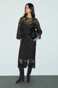Woman wearing a long, black lace dress with scalloped edges in a délicate motif fleuri, knee-high brown suede boots, and holding a black bag, standing against a plain light gray wall.