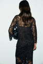 A woman with long brown hair, seen from behind, wears a black lace dress with a motif fleuri and holds a black handbag against a light background.