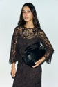 A woman in a black lace dress à motif fleuri holds a large black handbag. She has long dark hair and stands against a plain light background, looking directly at the camera with a neutral expression.