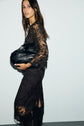 Woman in a long-sleeved, black lace top dentelle dress with a subtle motif fleuri, holding a large black bag, standing against a plain light-colored wall and looking slightly to the side.