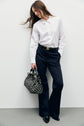 A woman stands against a white wall wearing a chemise blanche en popeline de coton, black trousers, black shoes, and holding a patterned black handbag.