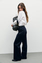 A person wearing a chemise blanche en popeline de coton and dark wide-leg pants stands on a gray floor, looking over their shoulder. They hold a black and gray patterned bag over one arm, with long hair loose over their back.