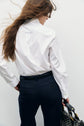 A person with long brown hair wears a chemise blanche en popeline de coton and navy trousers, seen from behind, holding a black patterned handbag in their right hand against a plain light background.