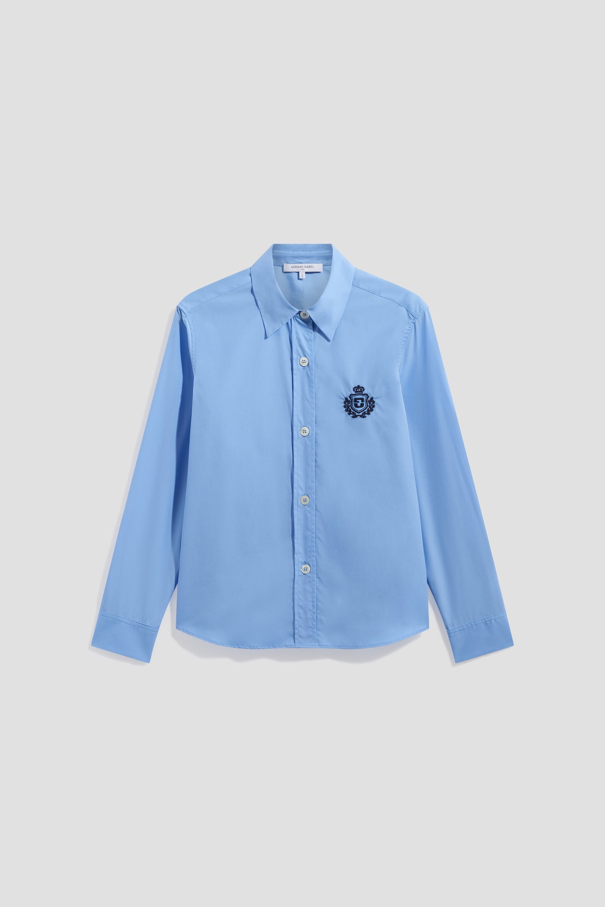 Light blue long-sleeve button-up shirt with a pointed collar and an embroidered dark blue crest (blason brodé) on the left chest, crafted from soft popeline de coton, displayed against a plain white background.