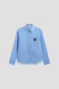 Light blue long-sleeve button-up shirt with a pointed collar and an embroidered dark blue crest (blason brodé) on the left chest, crafted from soft popeline de coton, displayed against a plain white background.