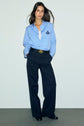 Woman stands against a plain background, wearing a chemise bleu ciel en popeline de coton avec un blason brodé, dark wide-leg trousers, a black belt with a gold buckle, and black shoes. She has long dark hair and a relaxed pose.