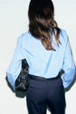 A person with long brown hair, wearing a chemise bleu ciel in popeline de coton and dark trousers, stands facing away and holds a black handbag under one arm.