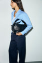 A woman wearing a chemise bleu ciel en popeline de coton and navy pants stands sideways, holding a black leather handbag under her arm against a plain light background.