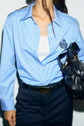 A person wearing a chemise bleu ciel en popeline de coton with a blason brodé, a white top underneath, dark pants with a belt, and holding a black bag. The head is mostly out of frame.