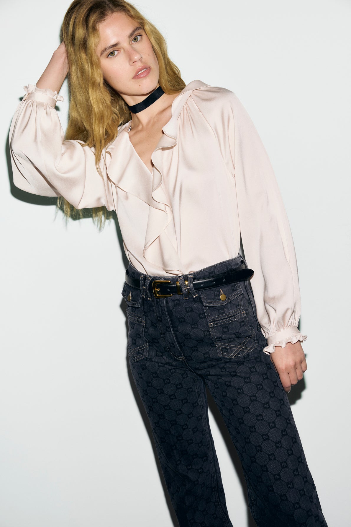 A person with long blonde hair wears a light col V à volants blouse with manches longues bouffantes, dark patterned high-waisted jeans, a black choker, and poses with one hand in their hair against a plain white background.