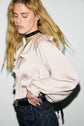 A person with long blonde hair wearing a chemise satinée à col V à volants and dark patterned jeans, accentuated by manches longues bouffantes and a black choker, poses against a light background with eyes closed, one hand resting on their hip.