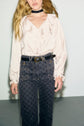 Woman wearing a chemise satinée à col V à volants with manches longues bouffantes, paired with high-waisted patterned jeans, a black belt, and a black choker, standing against a plain wall. Her face is partially out of the frame.