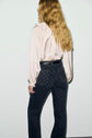 A person with long wavy blonde hair, wearing a pale pink chemise satinée à col V à volants and high-waisted black patterned jeans, stands facing away from the camera against a plain light background.