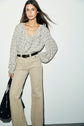 A woman stands against a white wall wearing a chemise à volants à carreaux, high-waisted beige pants with a black belt, and holding a black handbag.