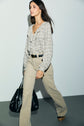 A person walks confidently wearing a beige chemise à carreaux and tan pants, holding a large black bag. The background is plain and well-lit.