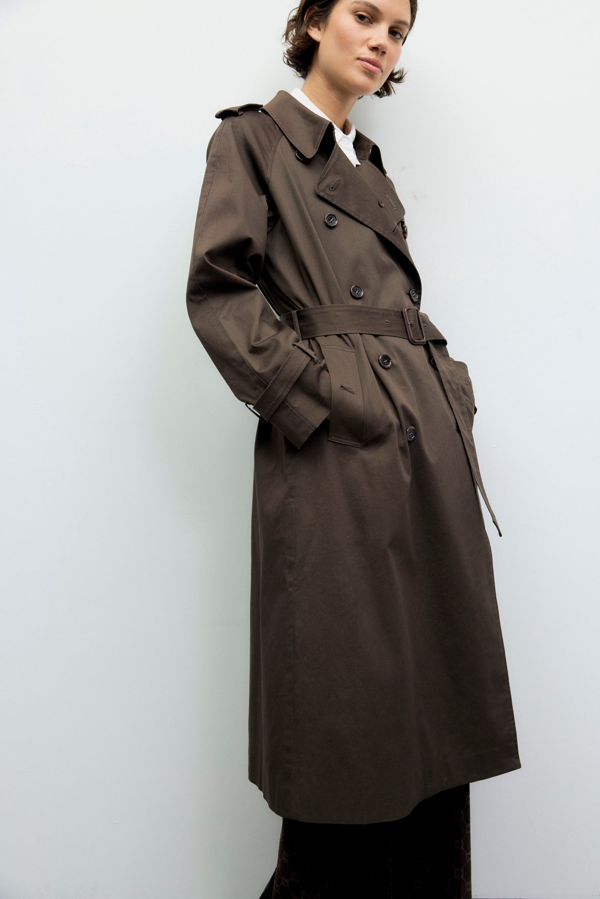 A person wearing a long, dark trench coat stands against a plain light background, looking confidently at the camera with hands in the manteau femme’s pockets.