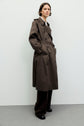 A person stands against a plain light wall, wearing a trench long, dark brown trench marron over trousers and black shoes. They have short, dark hair and a neutral expression, presenting an understated yet classic manteau femme look.