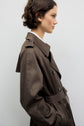 Woman with short brown hair wearing a manteau femme, a dark brown trench long coat, standing in profile against a plain light background.