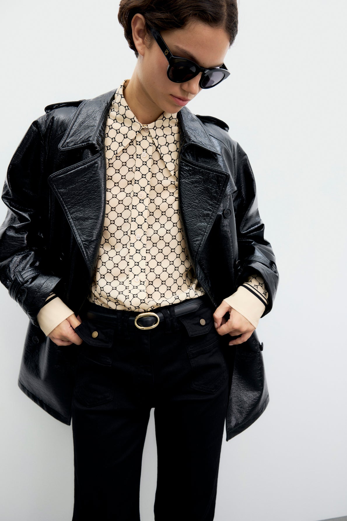 Person wearing sunglasses, a shiny black trench court, a patterned cream shirt, and black pants with a belt, standing against a plain white background.