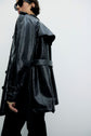 Person wearing a shiny black trench coat with a vernis craquelé noir finish, black pants, and sunglasses, facing sideways against a plain light background. The coat features a belted waist and textured finish, adding a veste tendance flair to the look.