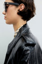 Person in profile wearing dark sunglasses, a shiny black vernis craquelé noir leather jacket, and a patterned high-collar shirt. The background is plain and light-colored.
